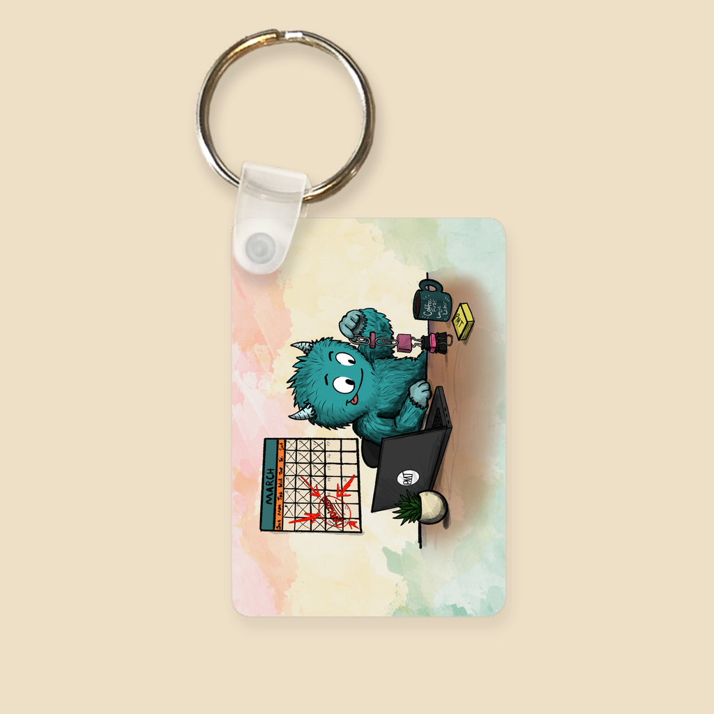 Keychain featuring a blue furry monster building a tower of office supplies beside a laptop while a calendar full of crossed-off days hangs in the background.