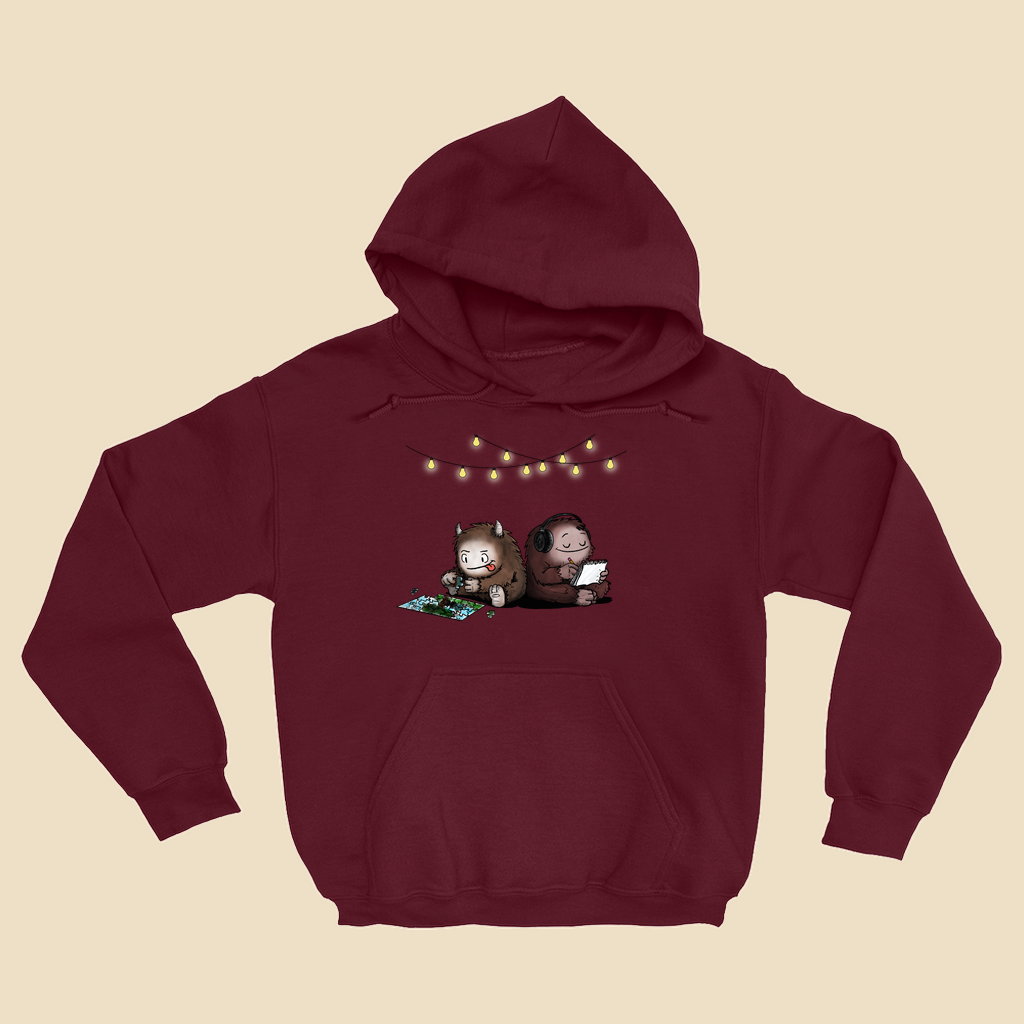 Hoodie featuring two cartoon creatures sitting back to back beneath string lights, one working on a puzzle and the other writing in a notebook.
