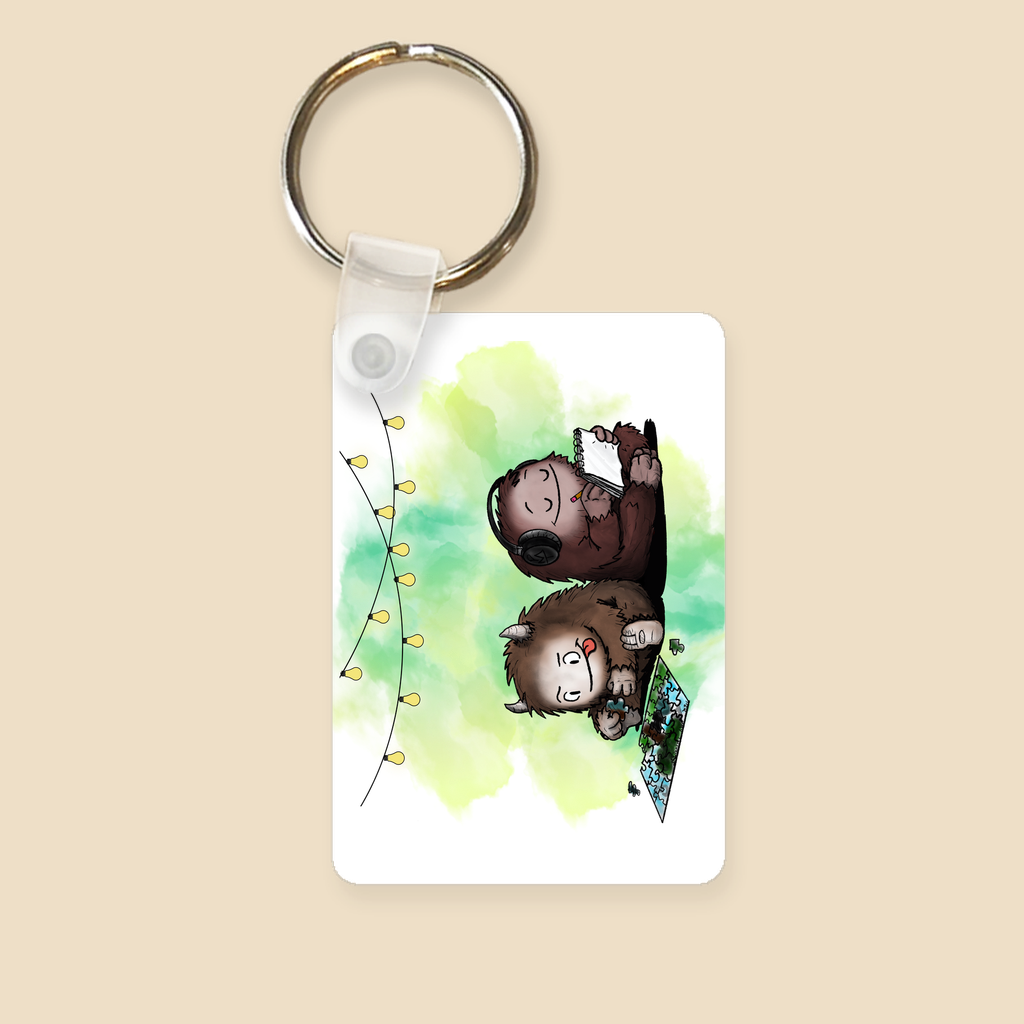 Keychain featuring two cartoon creatures sitting back to back beneath string lights, one working on a puzzle and the other writing in a notebook, with a warm watercolor background.