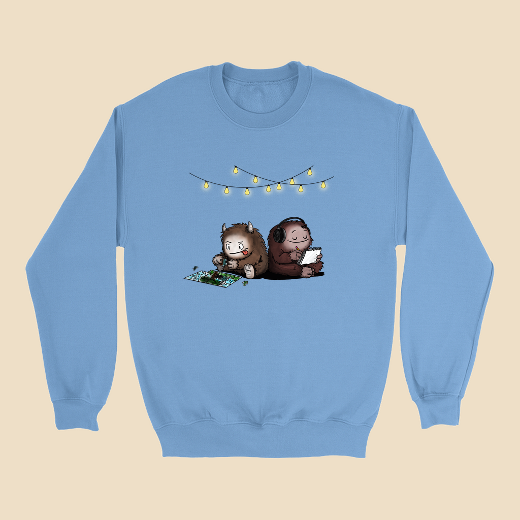 Sweatshirt featuring two cartoon creatures sitting back to back beneath string lights, one working on a puzzle and the other writing in a notebook.