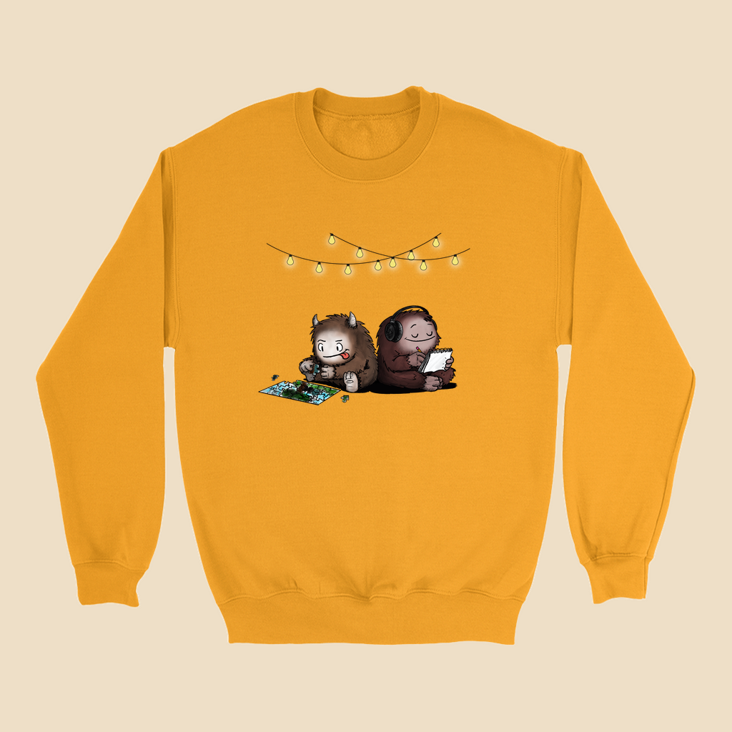 Sweatshirt featuring two cartoon creatures sitting back to back beneath string lights, one working on a puzzle and the other writing in a notebook.