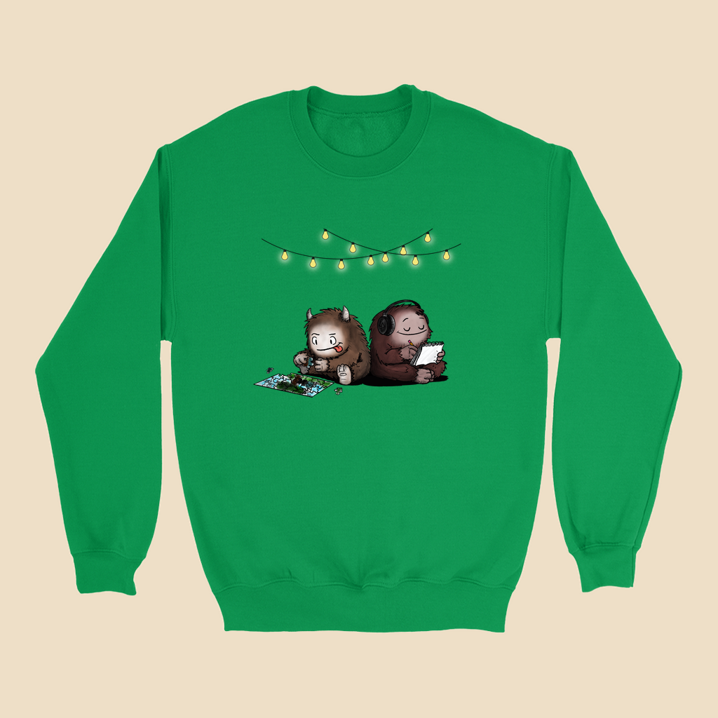 Sweatshirt featuring two cartoon creatures sitting back to back beneath string lights, one working on a puzzle and the other writing in a notebook.