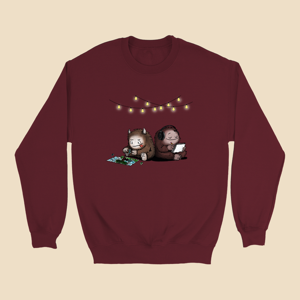 Sweatshirt featuring two cartoon creatures sitting back to back beneath string lights, one working on a puzzle and the other writing in a notebook.