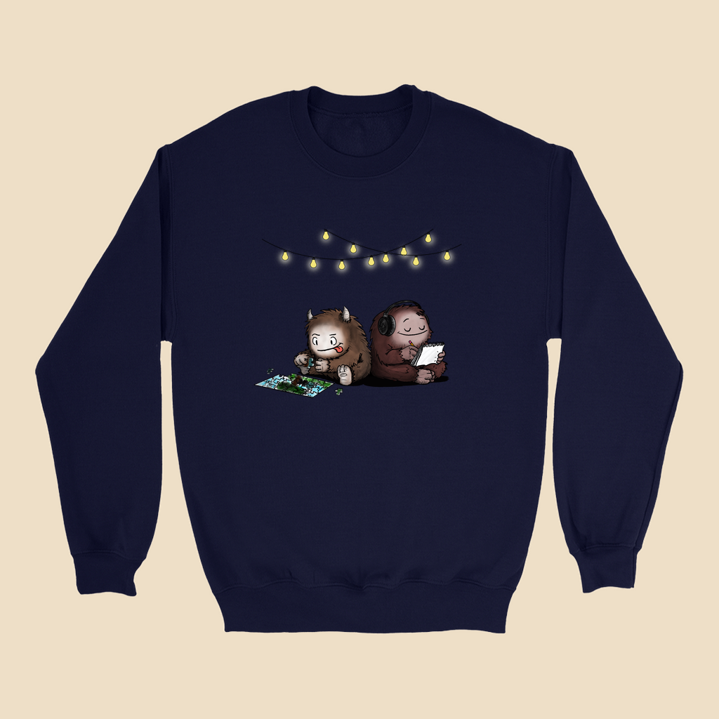 Sweatshirt featuring two cartoon creatures sitting back to back beneath string lights, one working on a puzzle and the other writing in a notebook.
