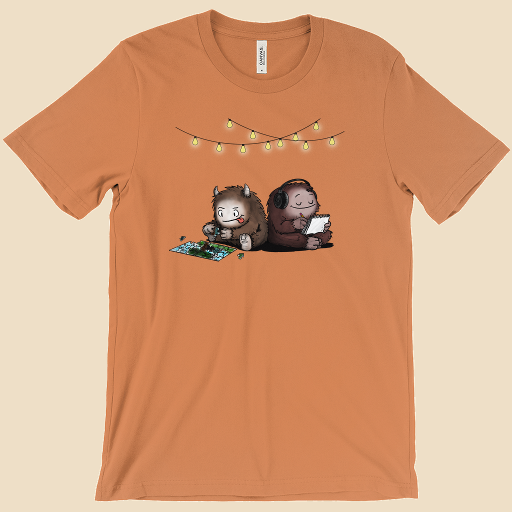 Parallel Play T-shirt featuring two cartoon creatures sitting back to back beneath string lights, one working on a puzzle and the other writing in a notebook.
