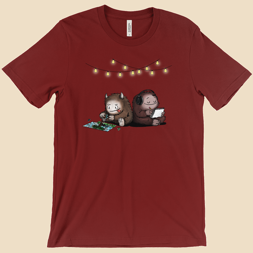 Parallel Play T-shirt featuring two cartoon creatures sitting back to back beneath string lights, one working on a puzzle and the other writing in a notebook.