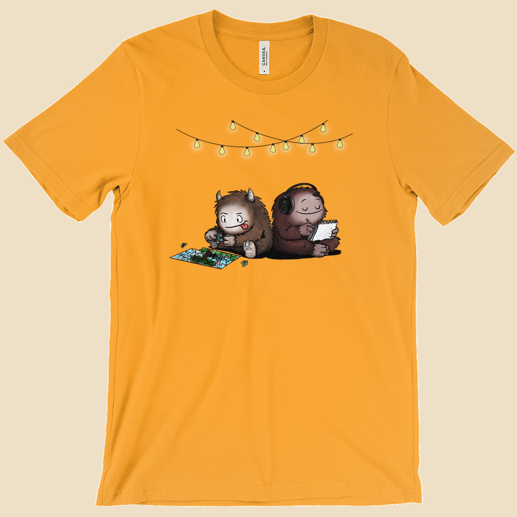 Parallel Play T-shirt featuring two cartoon creatures sitting back to back beneath string lights, one working on a puzzle and the other writing in a notebook.