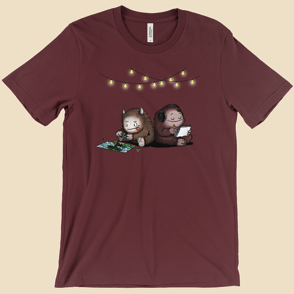 Parallel Play T-shirt featuring two cartoon creatures sitting back to back beneath string lights, one working on a puzzle and the other writing in a notebook.