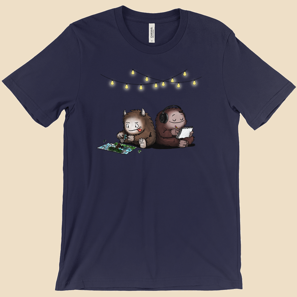 Parallel Play T-shirt featuring two cartoon creatures sitting back to back beneath string lights, one working on a puzzle and the other writing in a notebook.