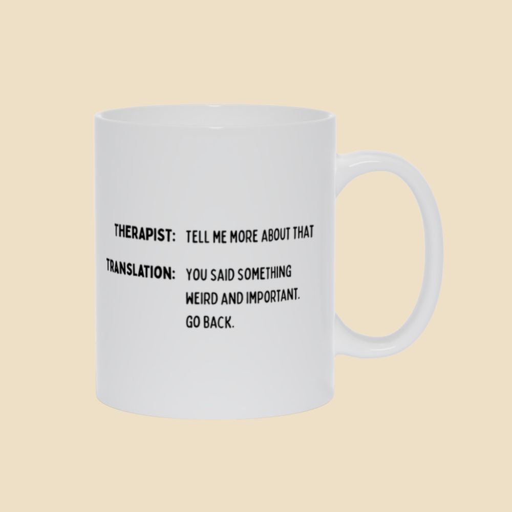 White mug with black text reading “Therapist: Tell me more about that. Translation: You said something weird and important. Go back.”