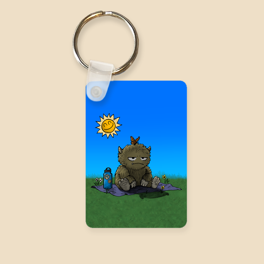Keychain featuring a furry monster sitting miserably in the sunshine while the cheerful world around it enjoys a beautiful day.